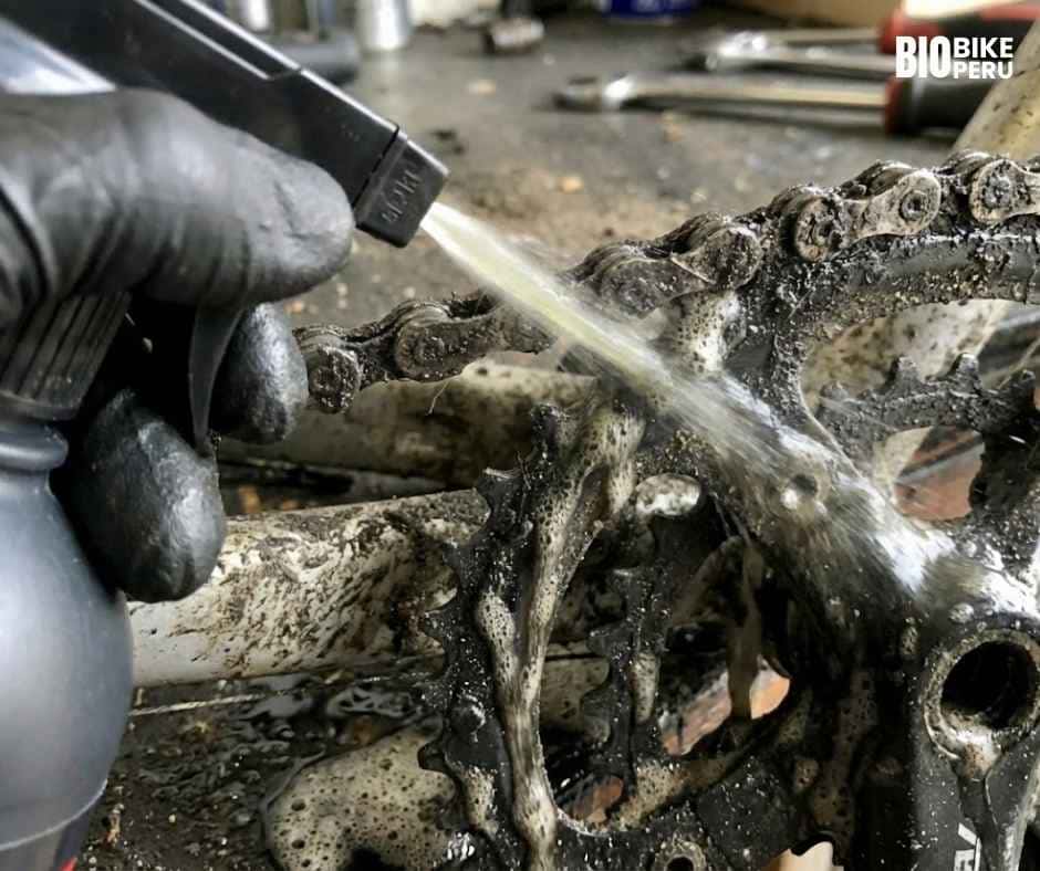 Primer plano de la limpieza de transmisión de bicicleta: mano con guante aplicando Biobike Desengrasante con una brocha sobre la cadena y el plato para remover la grasa acumulada.