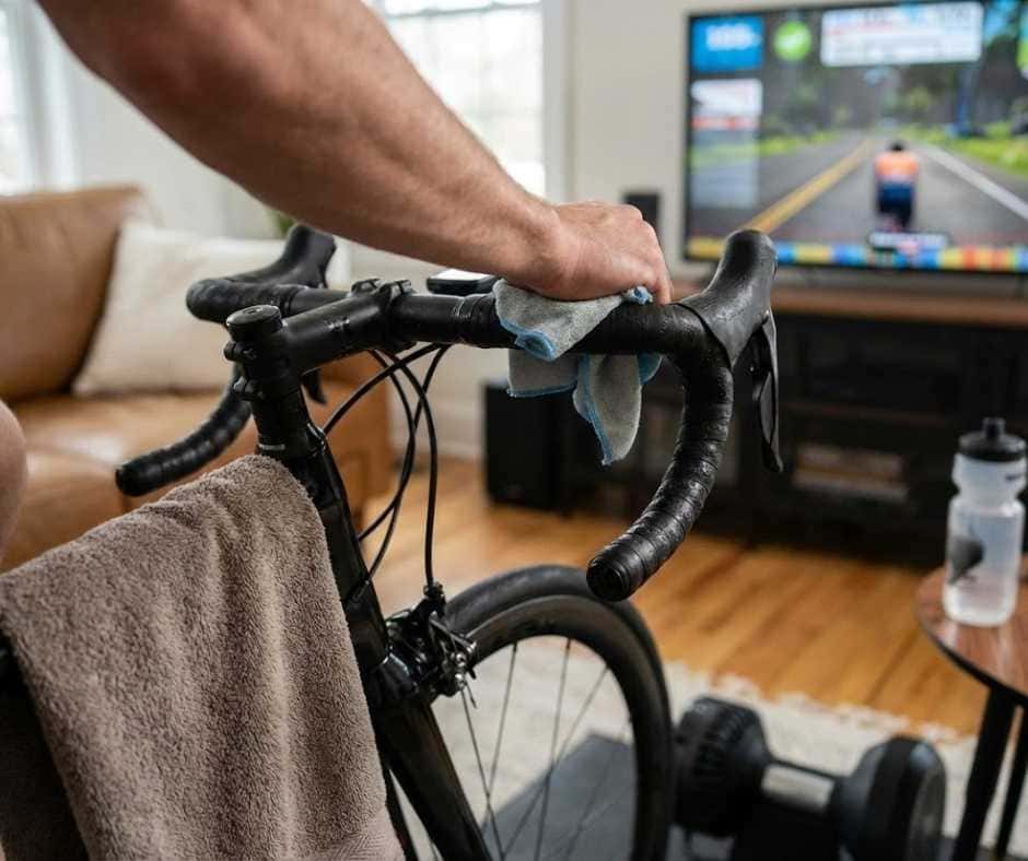 Ciclista limpiando el manillar y la potencia de su bicicleta con un paño de microfibra después de una sesión de ciclismo indoor, eliminando residuos de sudor para prevenir la corrosión.