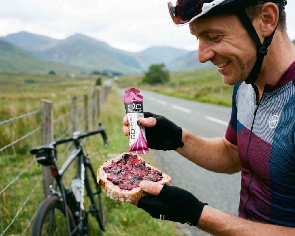 Ciclista en ruta sosteniendo un gel energético y un pastel de fruta, ilustrando el debate de nutrición deportiva: geles vs. comida real.